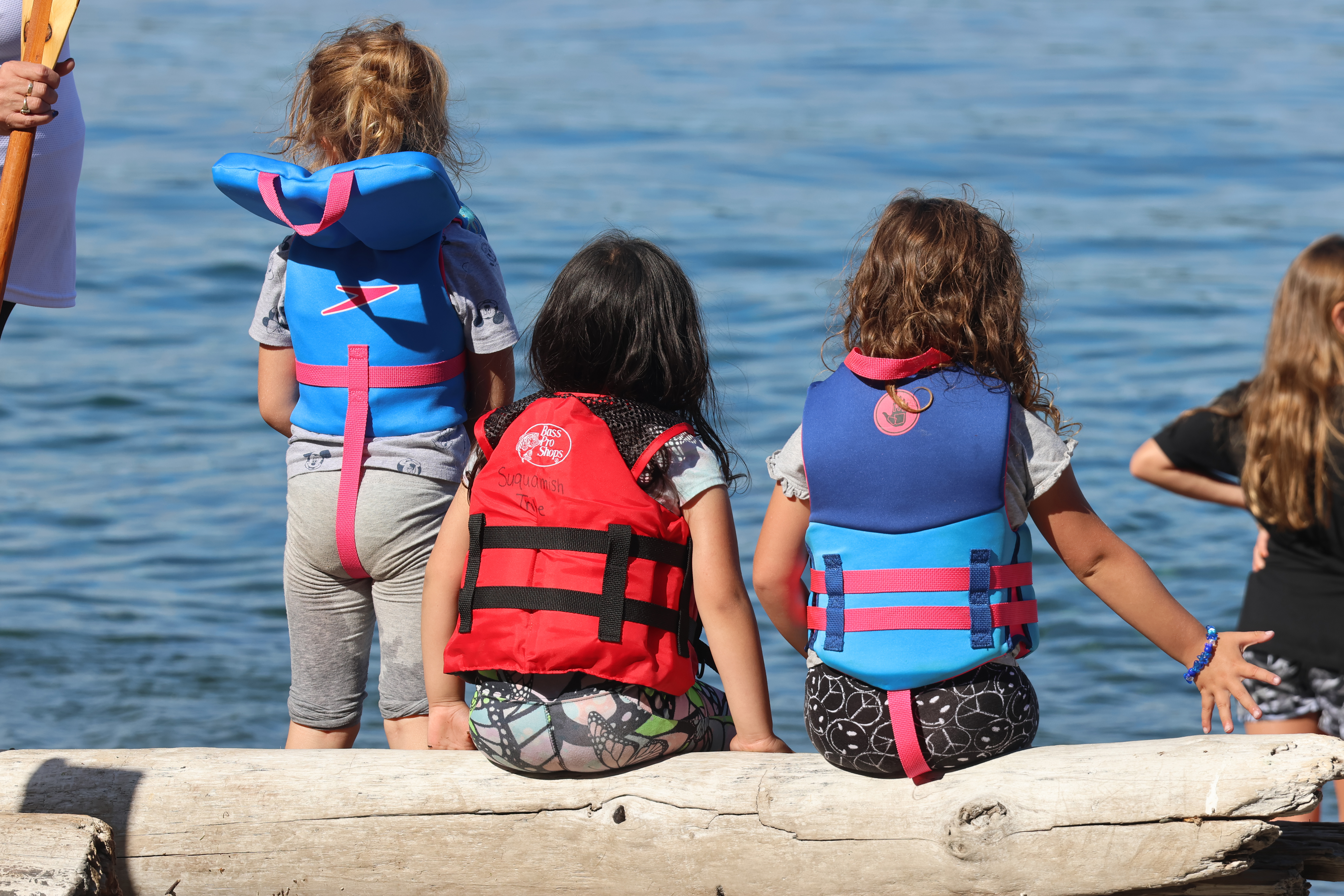 Three young girls rest on a log at Owen Beach on July 30, 2024. They watch as canoes land on their second to last day of the 2024 Canoe Journey.