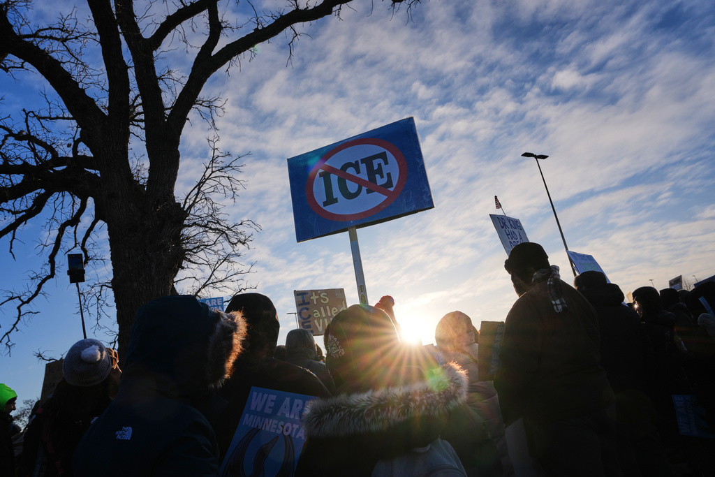 People gather for a protest. A person in the middle of the group holds a sign with the word "ICE" in a circle with a slash through it.
