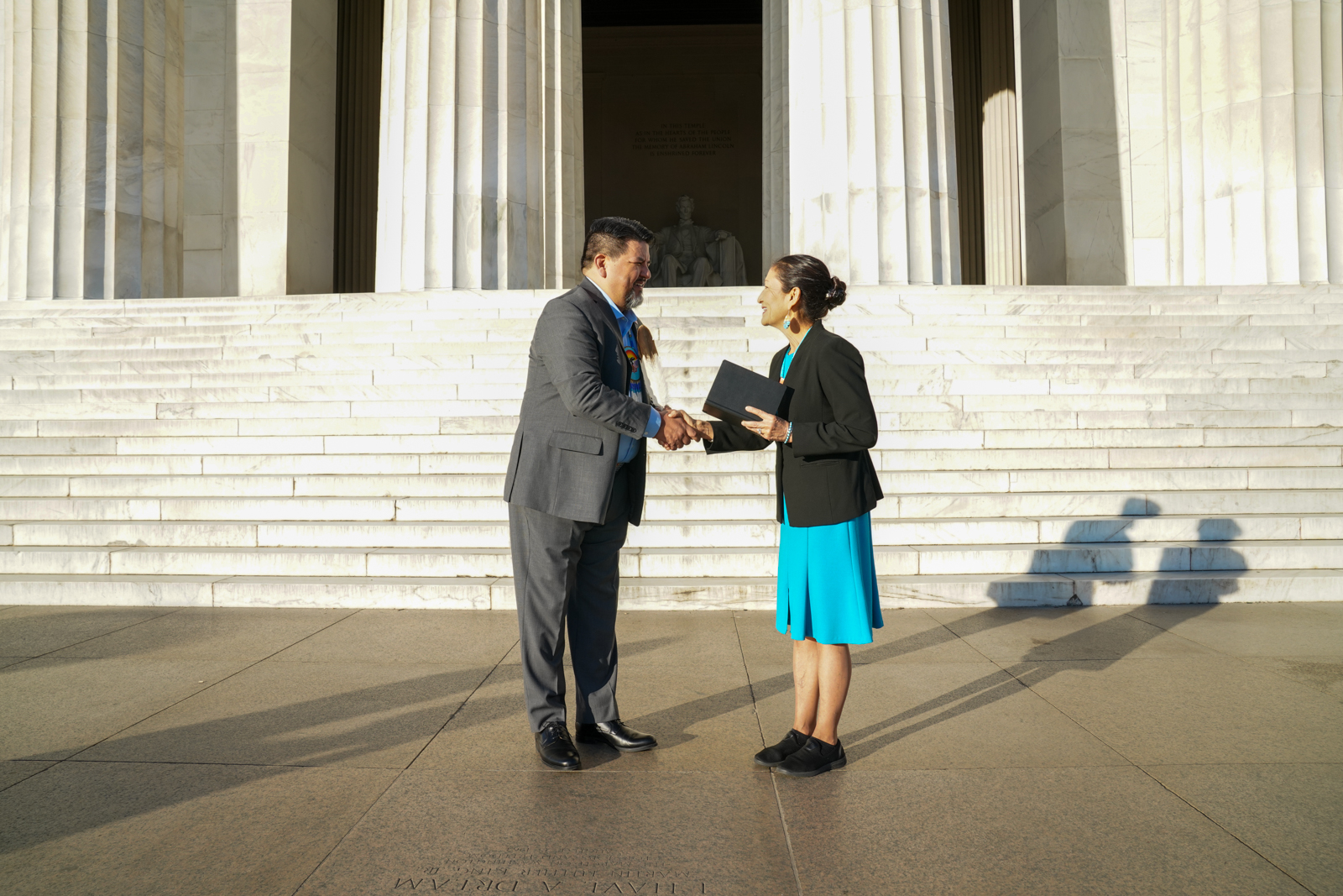 Sunlight colors the Lincoln Memorial steps light yellow. Charles Sams stands on the left in a dark gray suit and blue shirt, a beaded medallion hanging from a necklace on his chest and an eagle feather in his hair. Deb Haaland stands on the right in a turquoise dress, black blazer and turquoise jewelry. Each is smiling as they shake hands, Haaland holding a black book in her left hand. The Abraham Lincoln statue is covered in shadow and visible in the background.