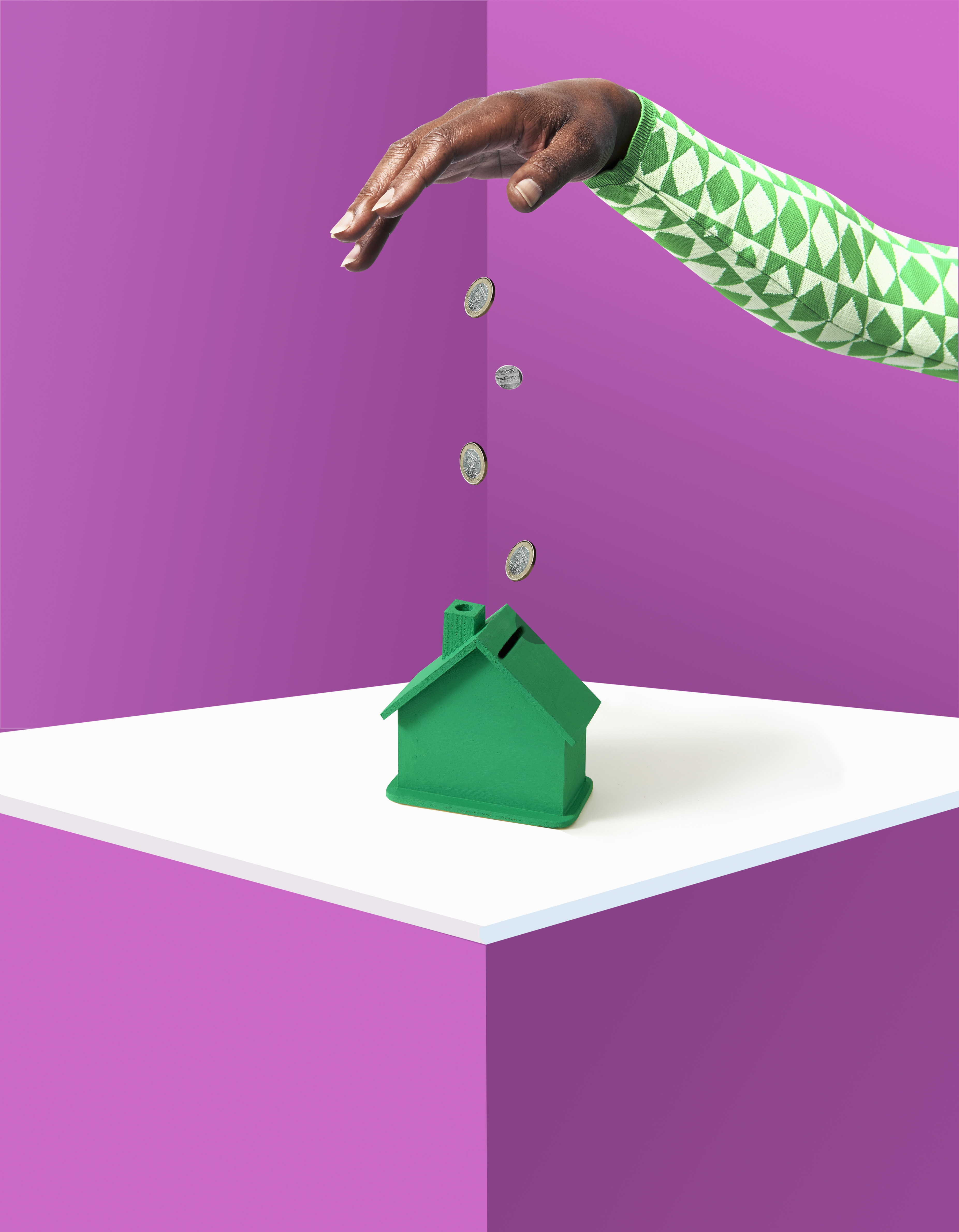 Studio photo of a hand dropping pound coins into a tiny green piggy bank-shaped like a house.  