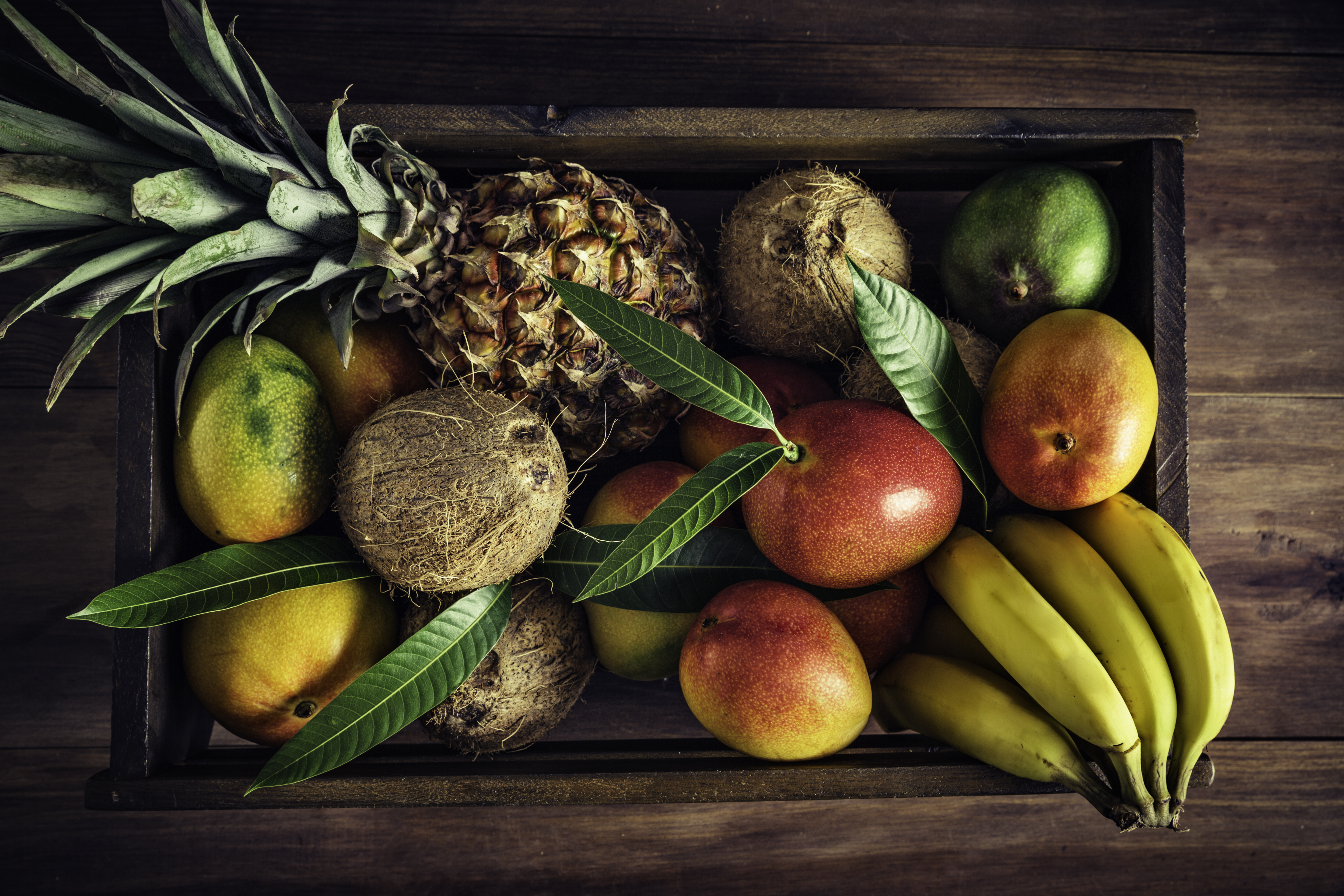 Wooden crates with assorted tropical fruits in rustic kitchen. Natural lighting. Mangos, pineapples, coconuts, bananas
