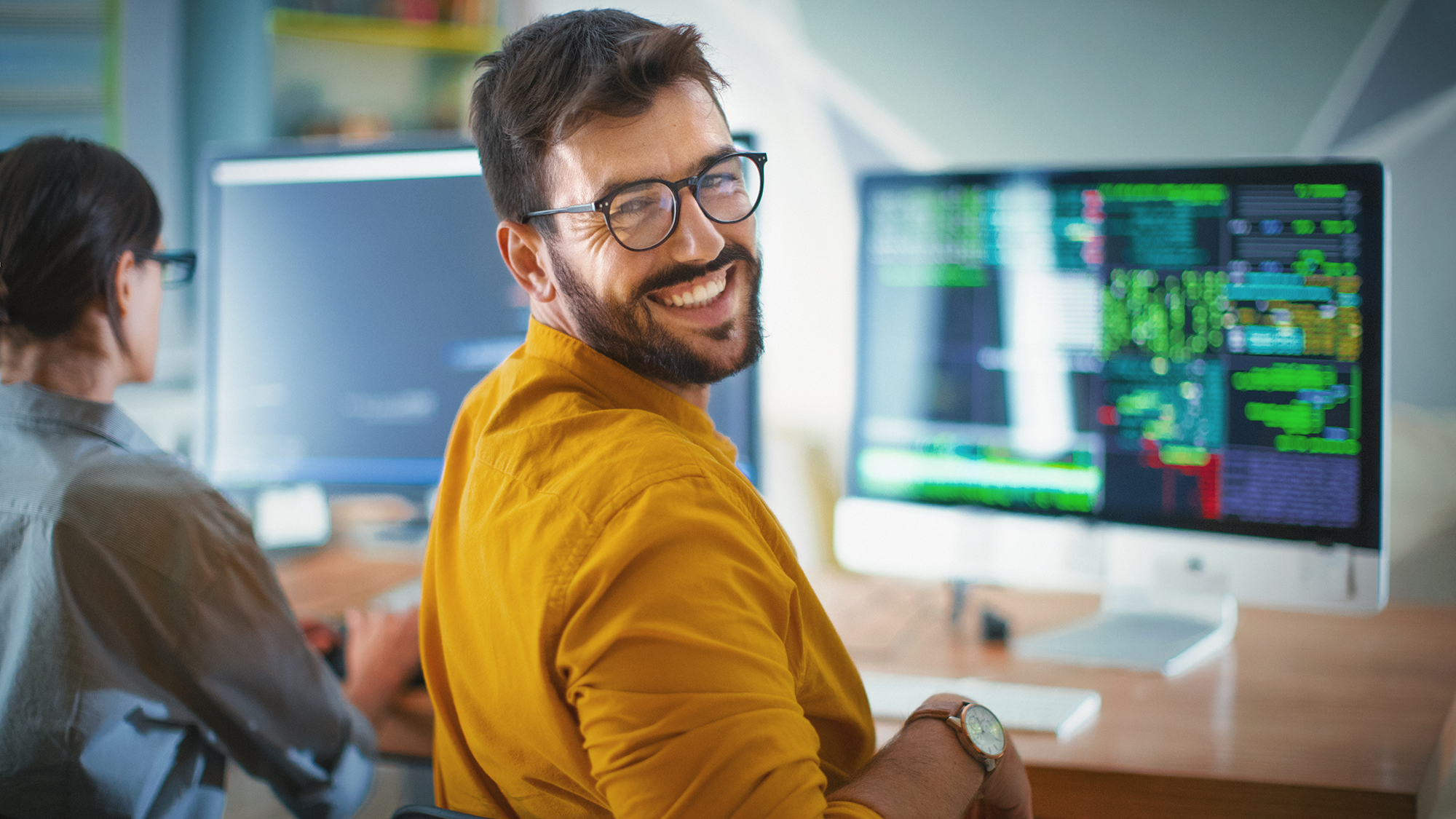 Primer plano sobre la vista del hombro de un alegre desarrollador de software de mediados de los años 20 en la oficina sonriendo a la cámara. Su compañera de trabajo está a su lado, todavía concentrada y trabajando.