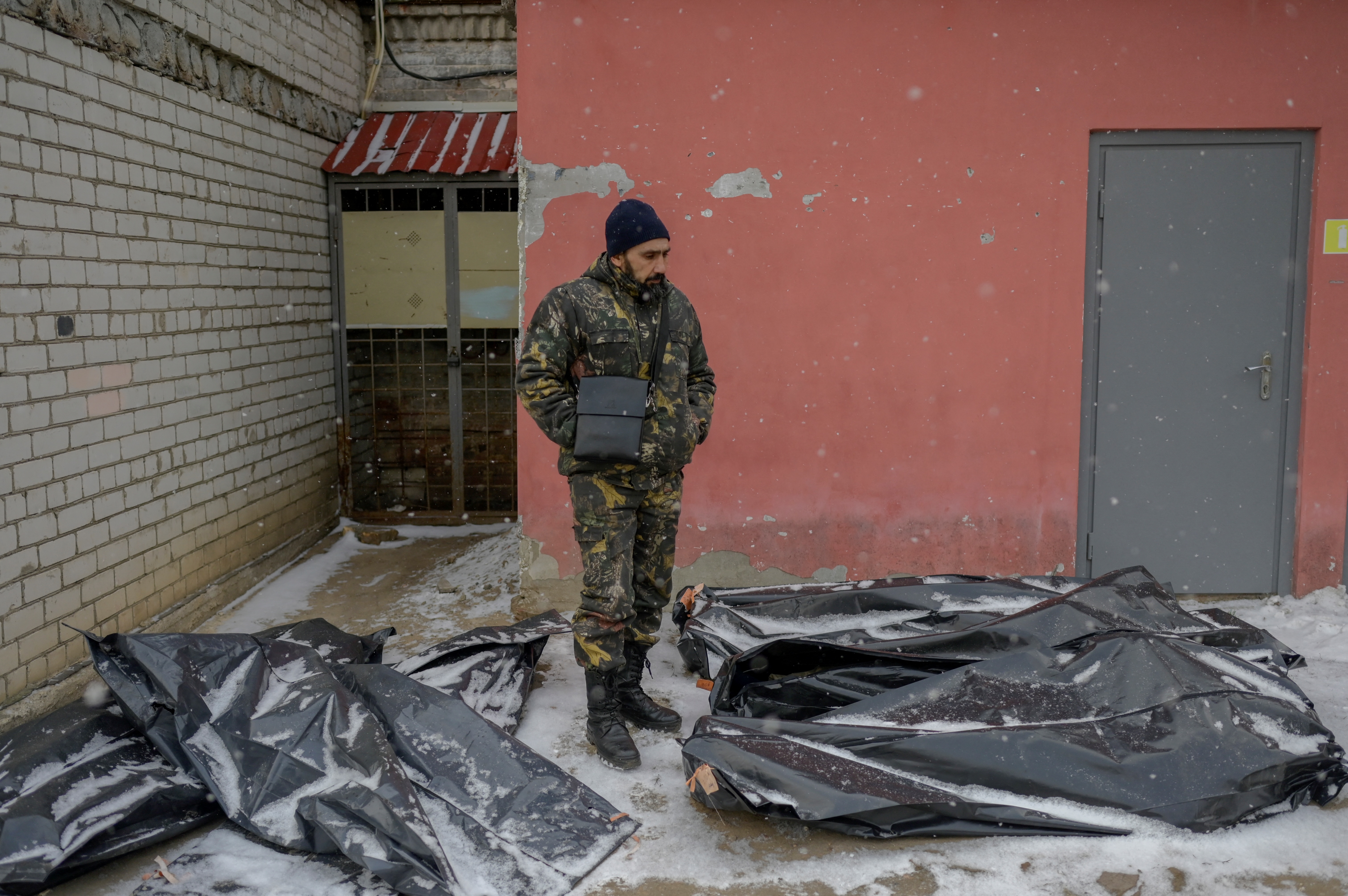 Un hombre ucraniano vestido de camuflaje se encuentra junto a bolsas para cadáveres cubiertas de nieve en el patio de una morgue en Mykolaiv, una ciudad a orillas del Mar Negro que ha estado bajo ataque ruso durante días el 11 de marzo de 2022. - El cadáver yace con su manos juntas, como en oración. De hecho, dice el empleado de la morgue, estaba lanzando cócteles molotov cuando los rusos lo atraparon. Le ataron las manos y lo ejecutaron. Tales son las escalofriantes imágenes de la morgue de Mykolaiv, una ciudad a orillas del Mar Negro que ha enfrentado días de ataque ruso. Afuera, en el patio del instituto forense donde se encuentra la morgue, la nieve cae constantemente sobre cadáveres envueltos en bolsas de plástico gris, que esperan ser evacuados. (Foto de BULENT KILIC / AFP)