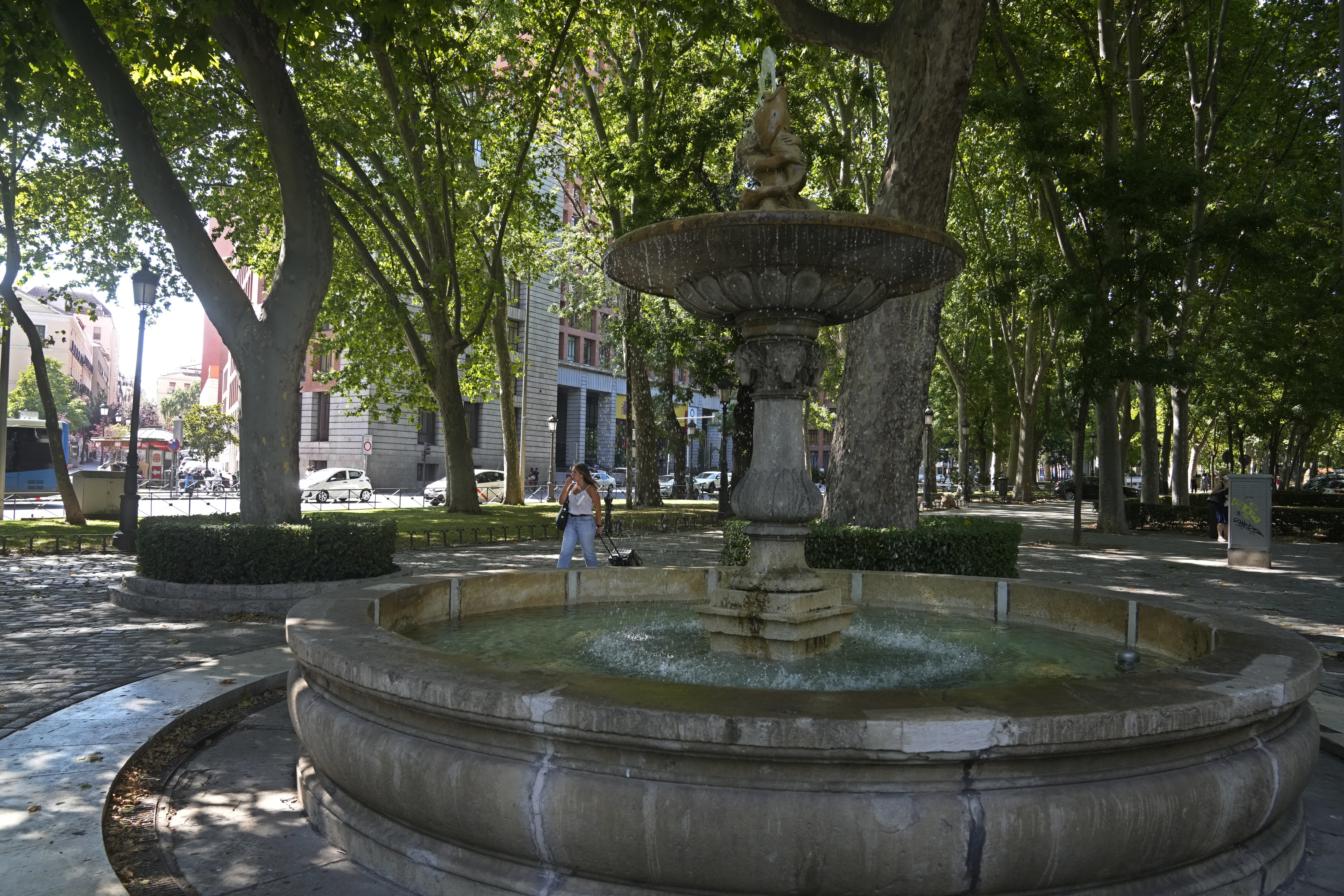 Una mujer camina junto a una fuente en el Paseo del Prado en Madrid, España, el viernes 23 de julio de 2021. El arbolado Paseo del Prado de Madrid y el parque del Retiro adyacente han sido incluidos en la lista del Patrimonio Mundial de la UNESCO. El Comité del Patrimonio Mundial de la UNESCO respaldó la candidatura que destaca la introducción de la naturaleza en la capital española. (AP Photo/Paul White)