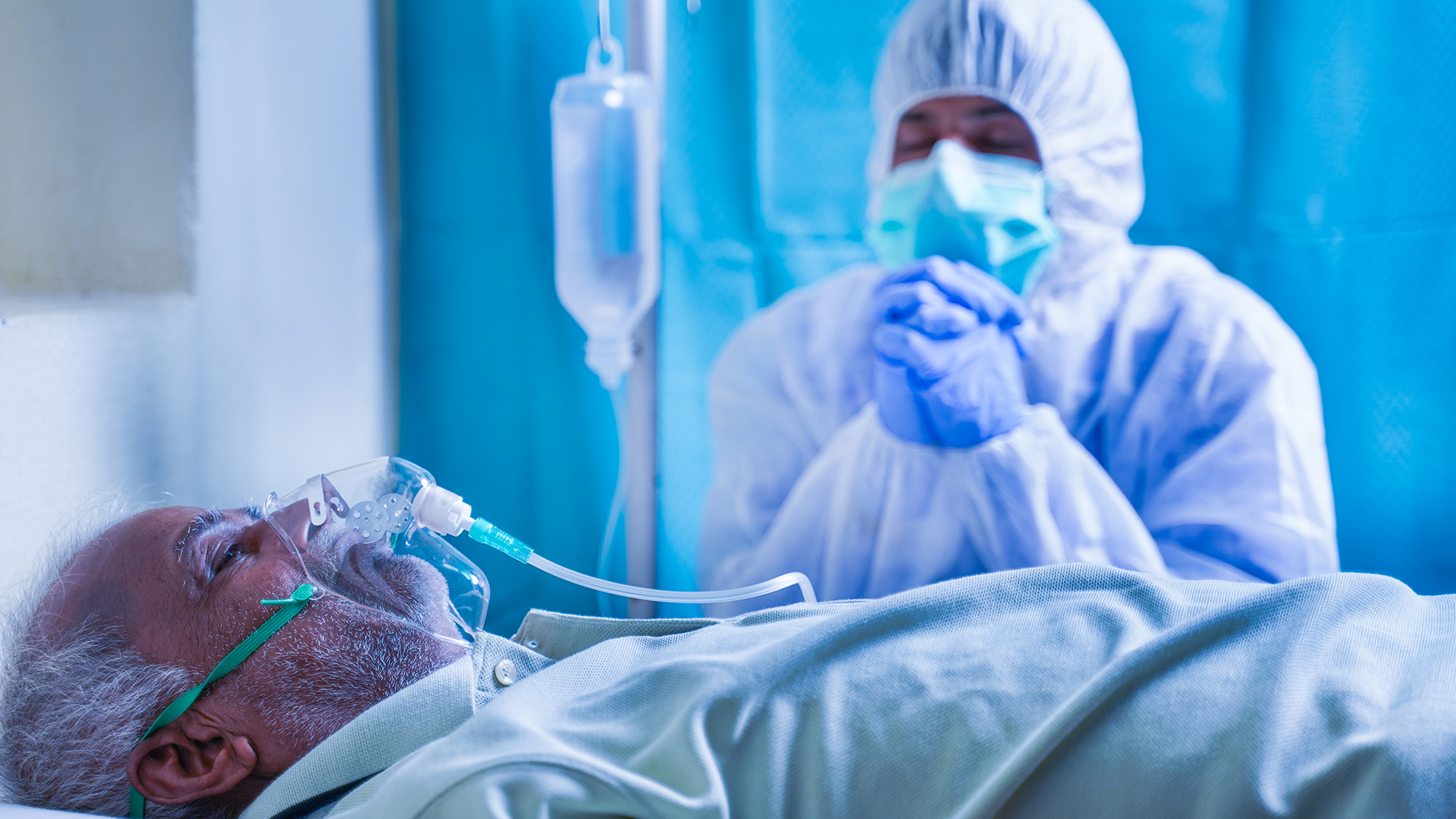 Preocupado miembro de la familia o médico con EPP o traje protector rezando por el coronavirus o el paciente infectado con covid-19 para recuperarse en el hospital mientras el paciente respira con una máscara de oxígeno del ventilador