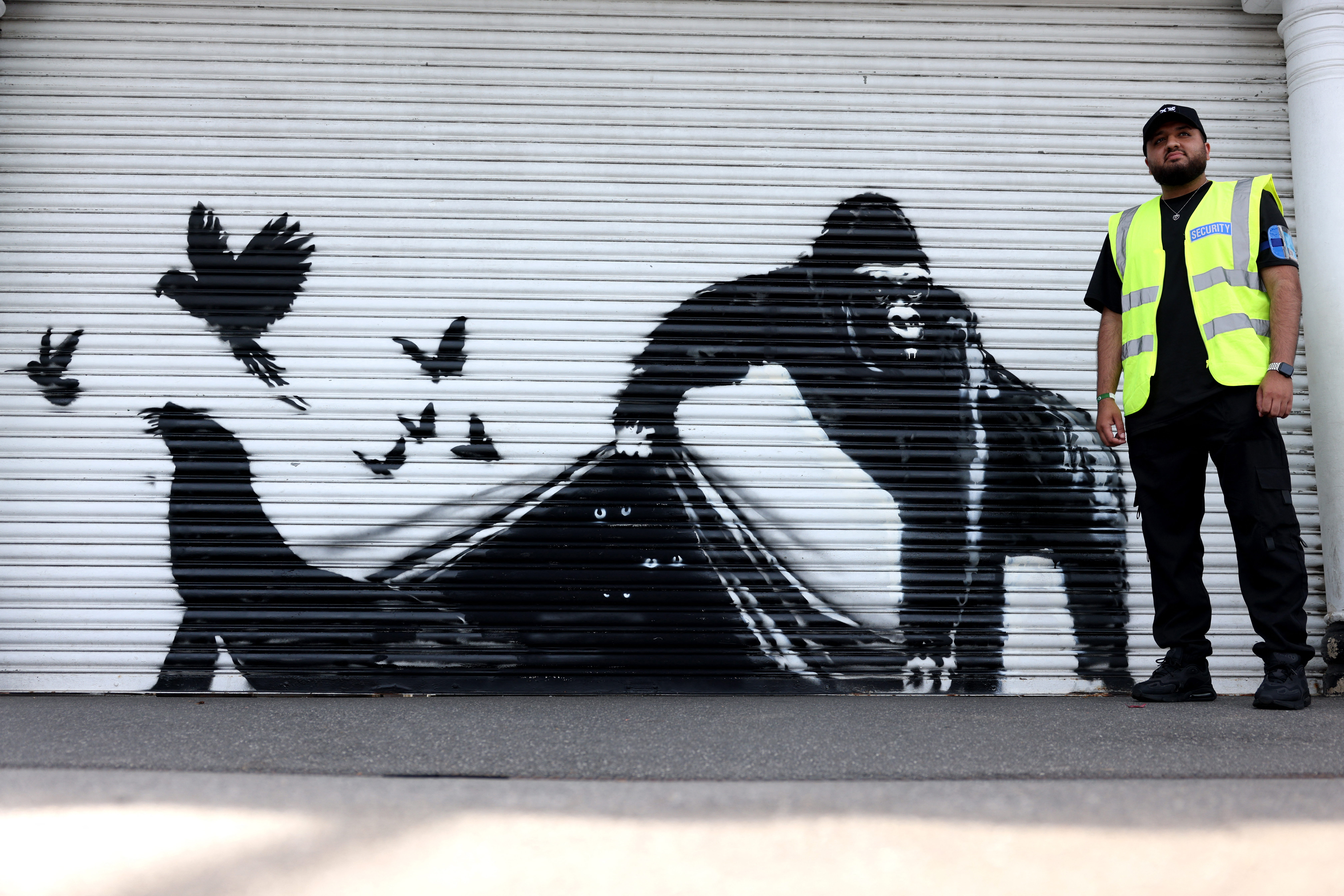 A security guard stands by an artwork by street artist Banksy, the ninth to released in nine days, depicting a gorilla releasing animals, on the front of a shutter outside London zoo, on August 13, 2024. The artist confirmed the work to be theirs after posting an image of it on the social media app Instagram. (Photo by Adrian DENNIS / AFP) / RESTRICTED TO EDITORIAL USE - MANDATORY MENTION OF THE ARTIST UPON PUBLICATION - TO ILLUSTRATE THE EVENT AS SPECIFIED IN THE CAPTION