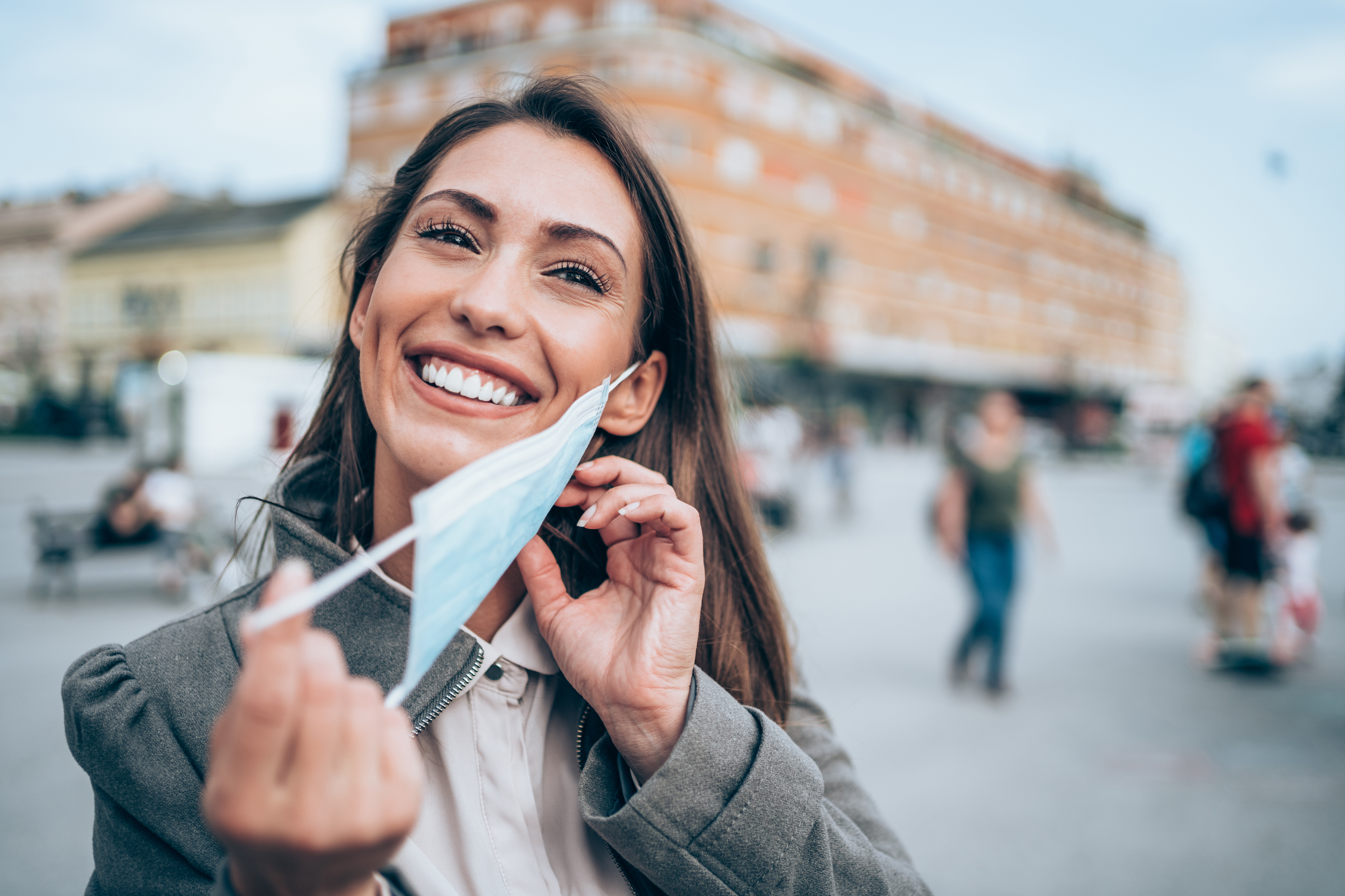 Hermosa joven feliz con máscara facial médica protectora para la protección contra virus durante la pandemia de Coronavirus/COVID-19. Mujer joven poniendo/quitando su máscara al aire libre en la ciudad.
