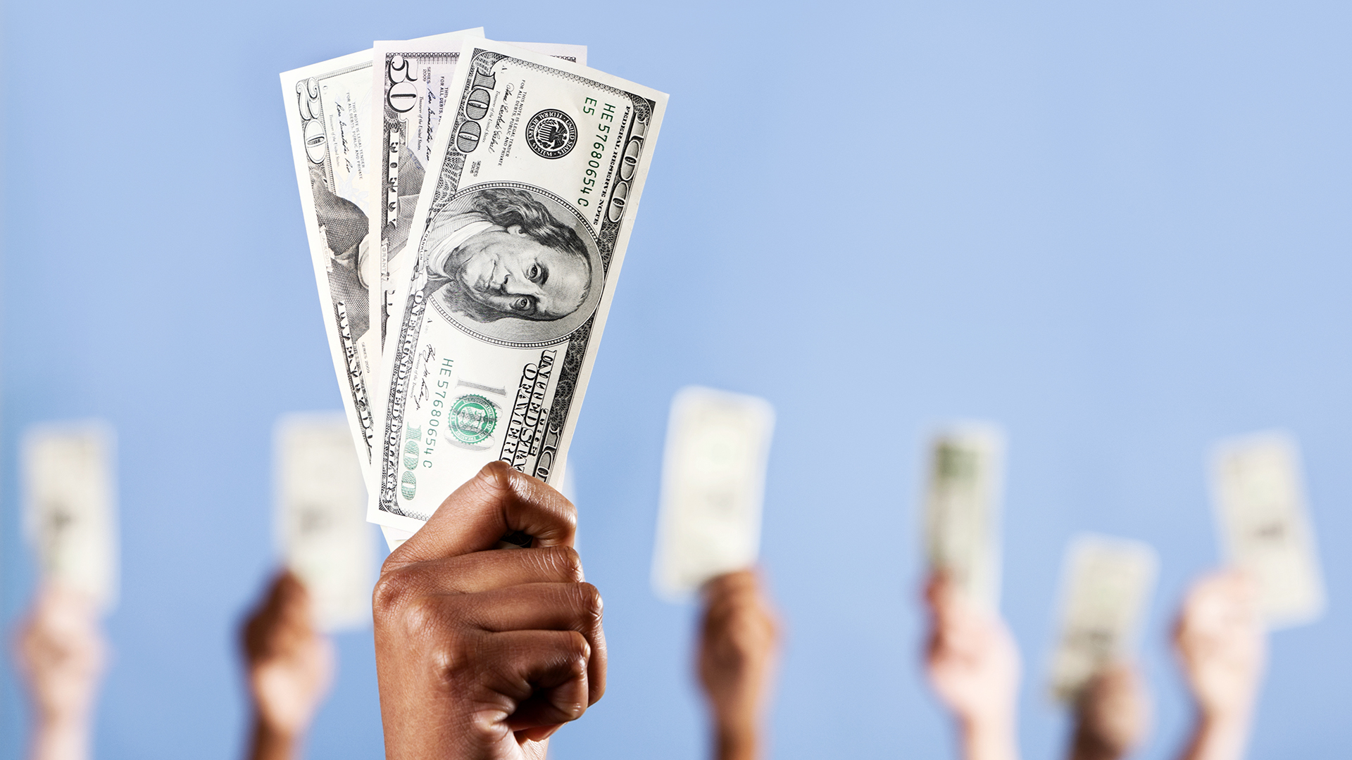 A group of hands of mixed sexes and races, all holding up US dollars. Focus is on the African man's hands in the foreground.