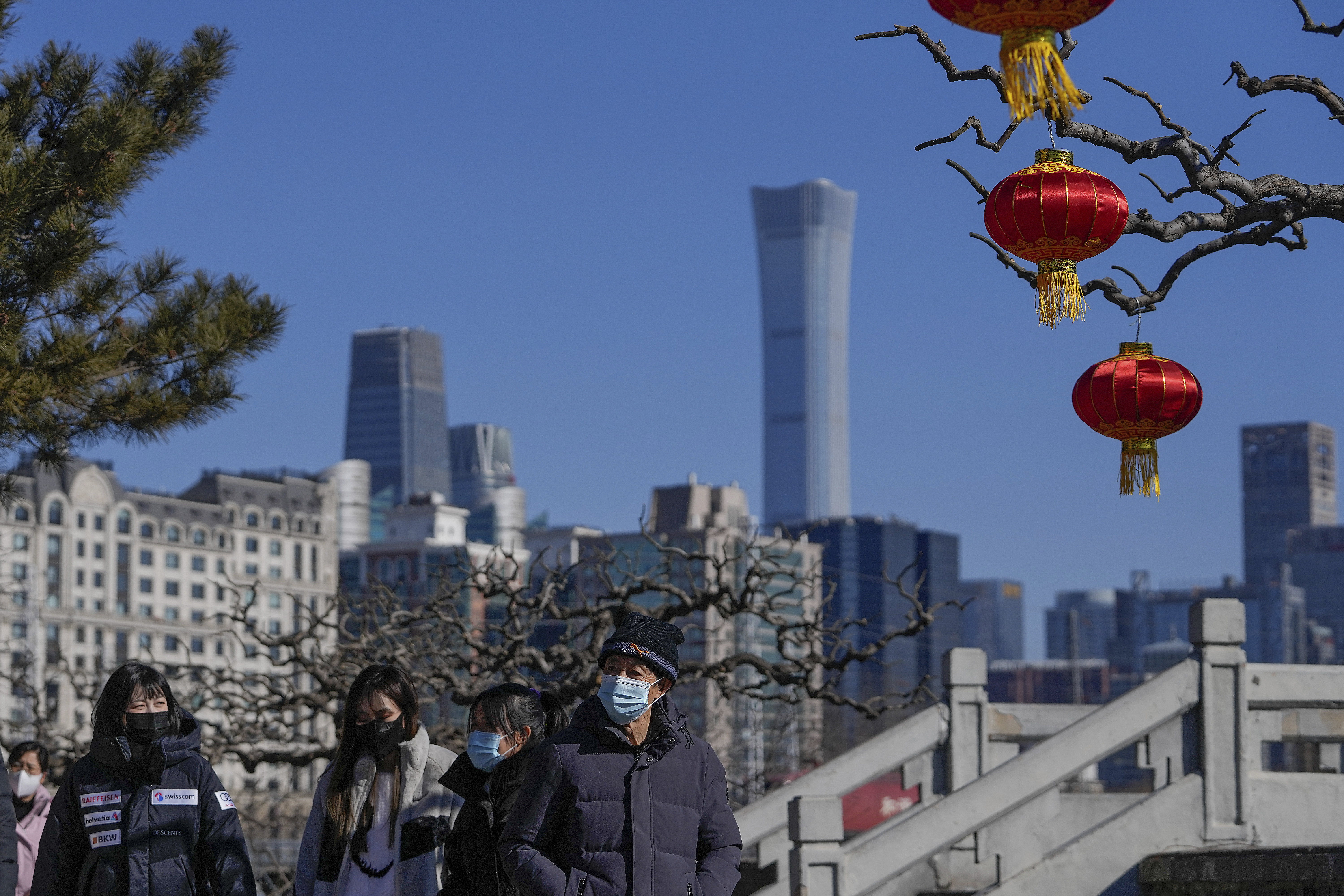 People wearing face masks to help protect from the coronavirus walk by lantern decorations for the upcoming Lunar New Year in a park seen against the city skyline in Beijing, Thursday, Jan. 27, 2022. (AP Photo/Andy Wong)