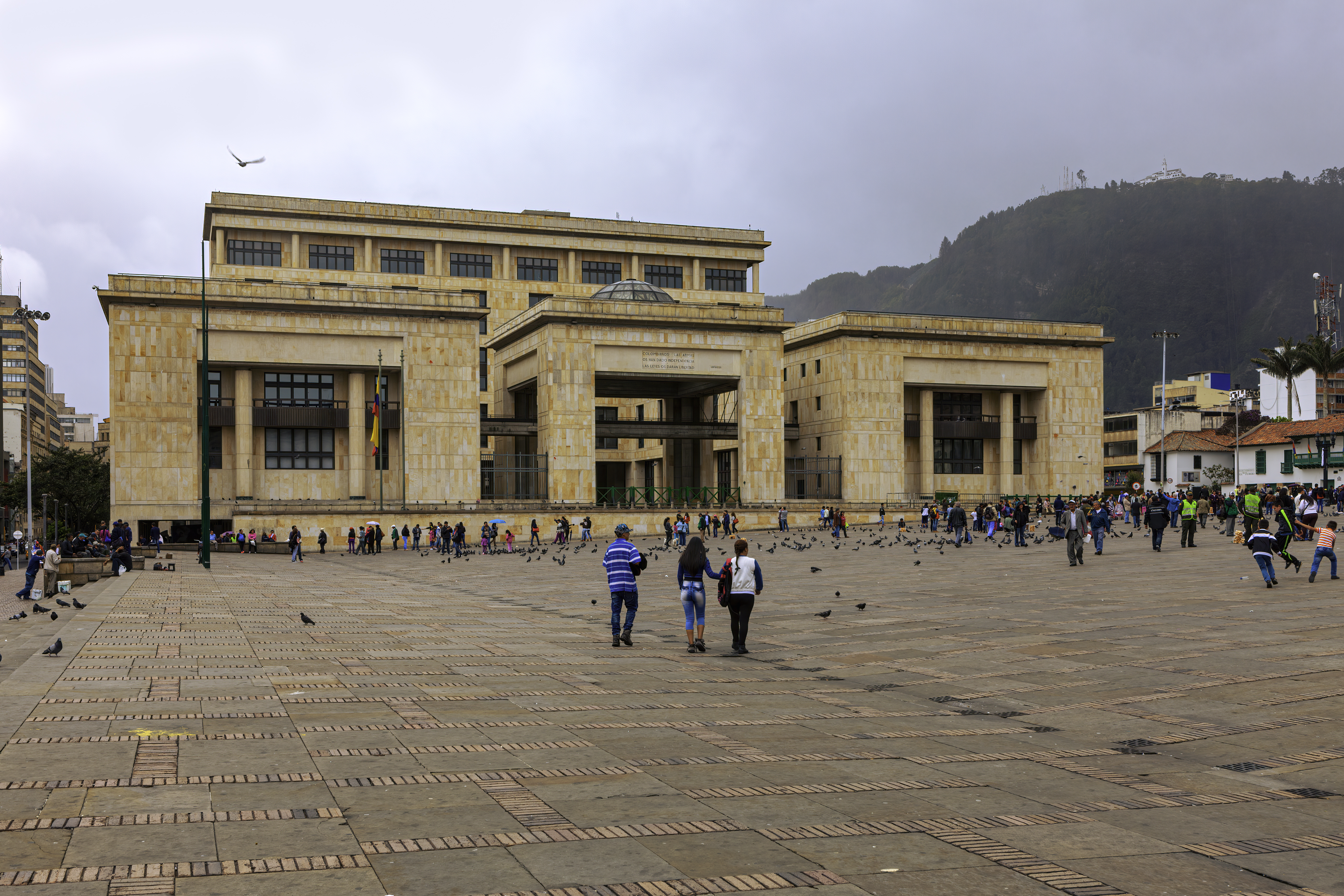 Mirando hacia la esquina noreste de la Plaza Bolívar en la capital andina de Bogotá en Colombia, Sur América. El edificio a la vista es el 'Palacio de Justicia' o traducido, el Palacio de Justicia. Es la Corte Suprema del País. Muchos colombianos locales pasan despreocupadamente, mientras realizan su día a día mientras conversan con amigos o familiares. Como muchas otras plazas en todo el mundo, hay muchas palomas en la plaza esperando a que los visitantes las alimenten. A la derecha de la imagen se puede ver el pico andino de Monserrate. El cielo está nublado; lloverá en breve. La altitud al nivel de la calle es de unos 8.500 pies sobre el nivel del mar. La mayoría de las personas usan una chaqueta o una capa adicional de ropa para mantenerse abrigados. Foto tomada a la luz del sol de la tarde en un día nublado; formato horizontal.