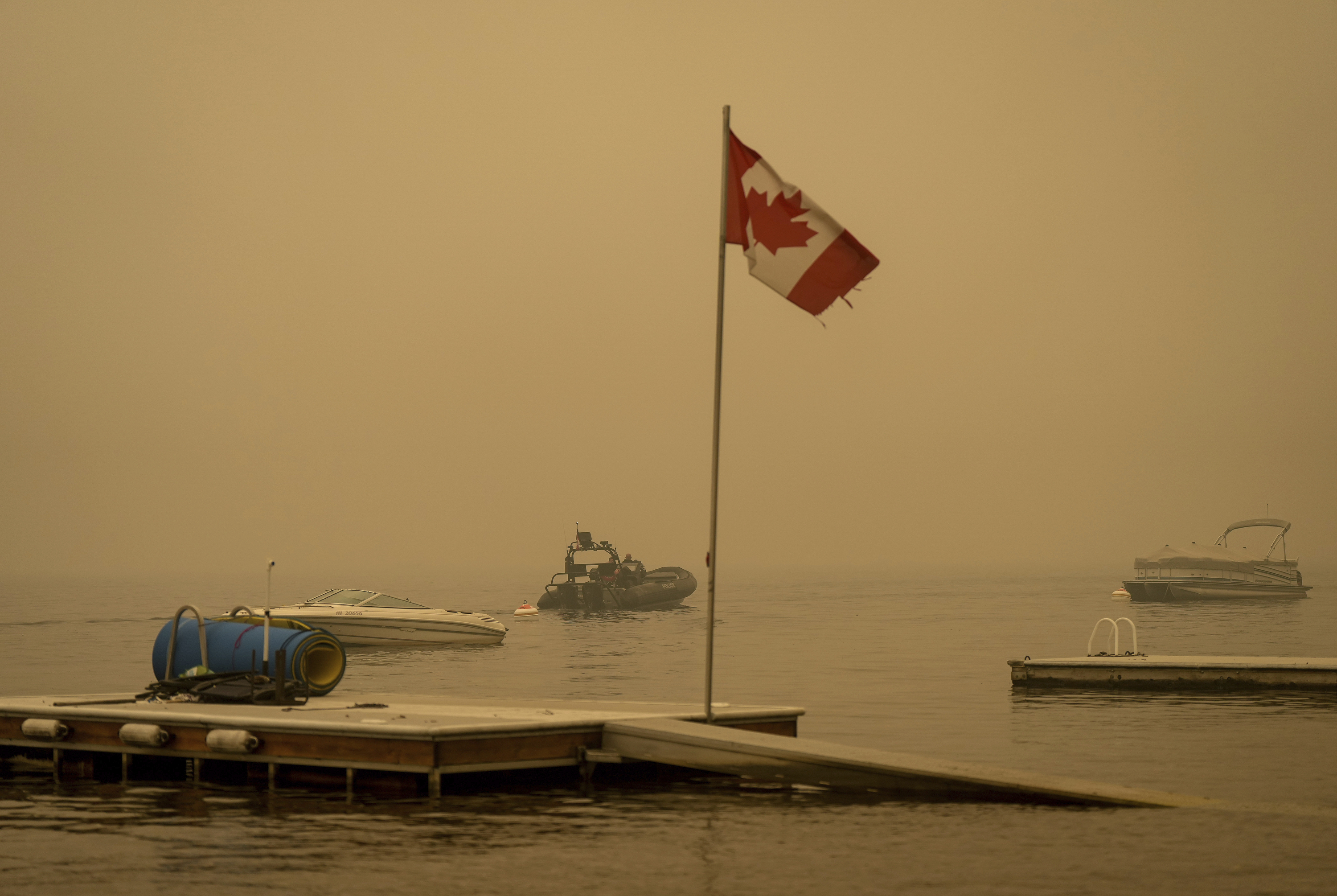 El humo espeso del incendio forestal del lago Lower East Adams llena el aire alrededor de una bandera canadiense ondeando en el viento mientras los oficiales de la Real Policía Montada de Canadá en un barco patrullan el lago Shuswap, en Scotch Creek, Columbia Británica, el domingo 20 de agosto de 2023