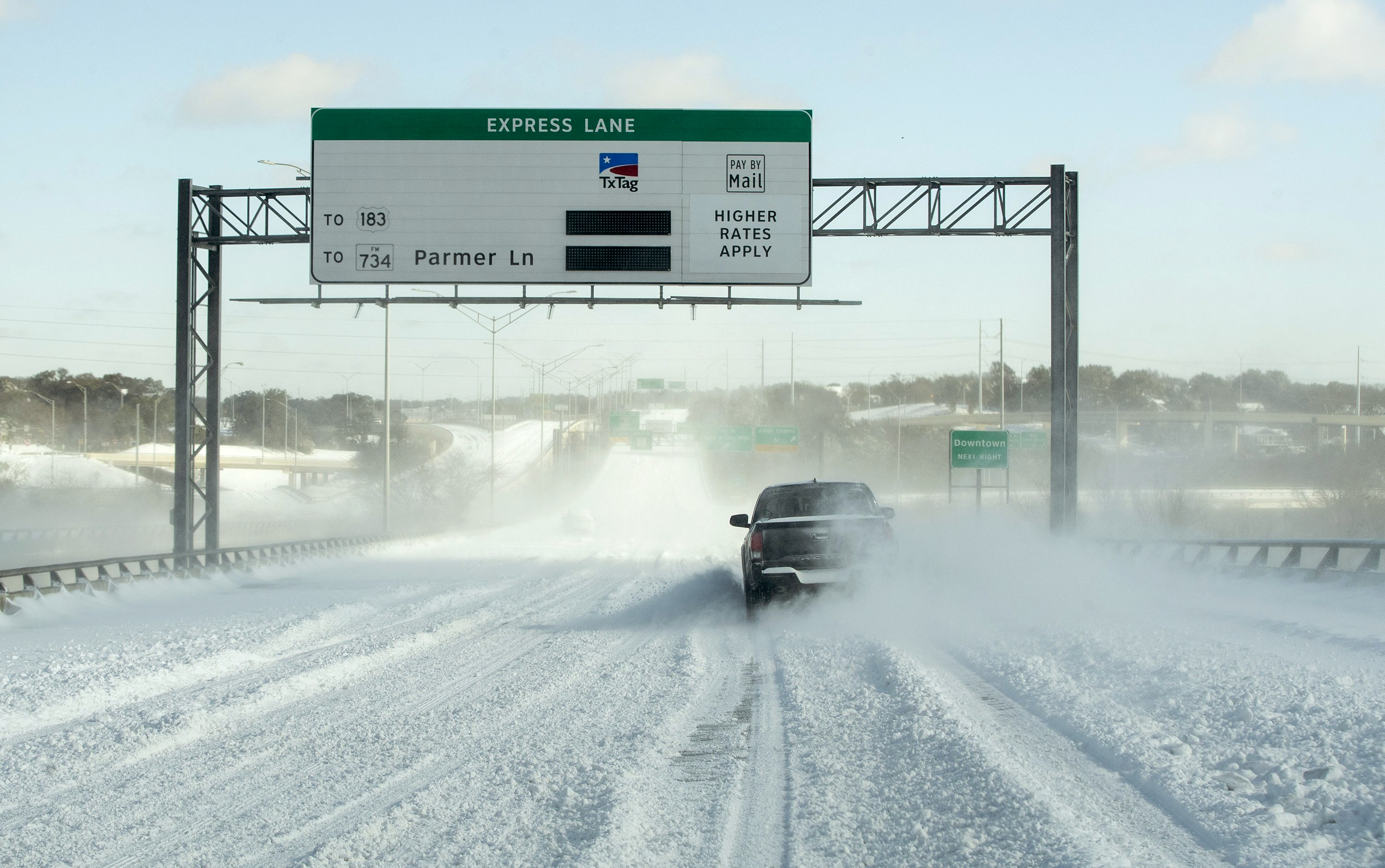 A truck drives in the snow on a Texas highway. A toll road saying "Express Lane to 183 to 737 Parmer Ln" appears above the road with its power out.