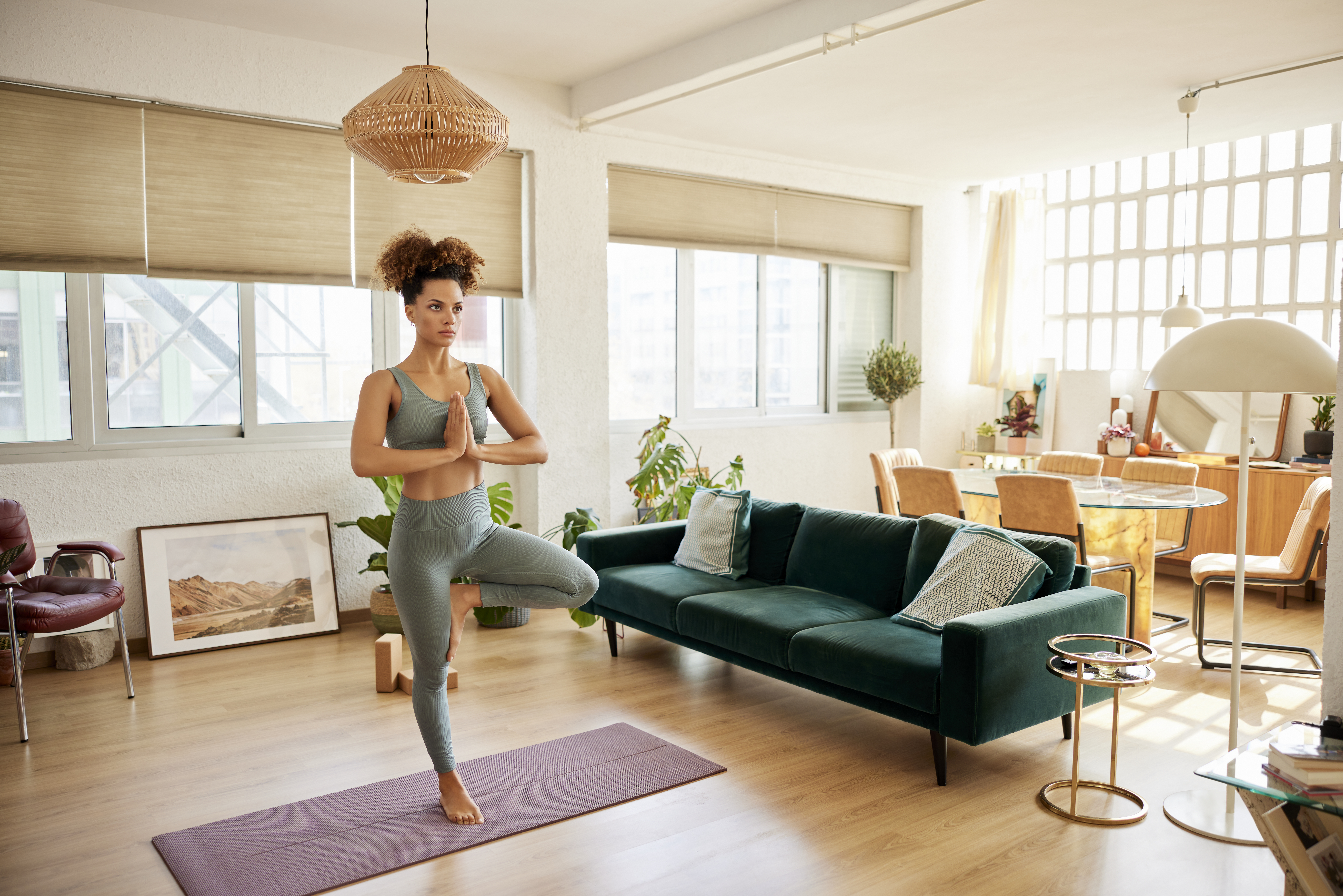 Full length of fit woman practicing tree pose at home. Young woman standing on one leg on exercise mat. She is doing yoga in living room.