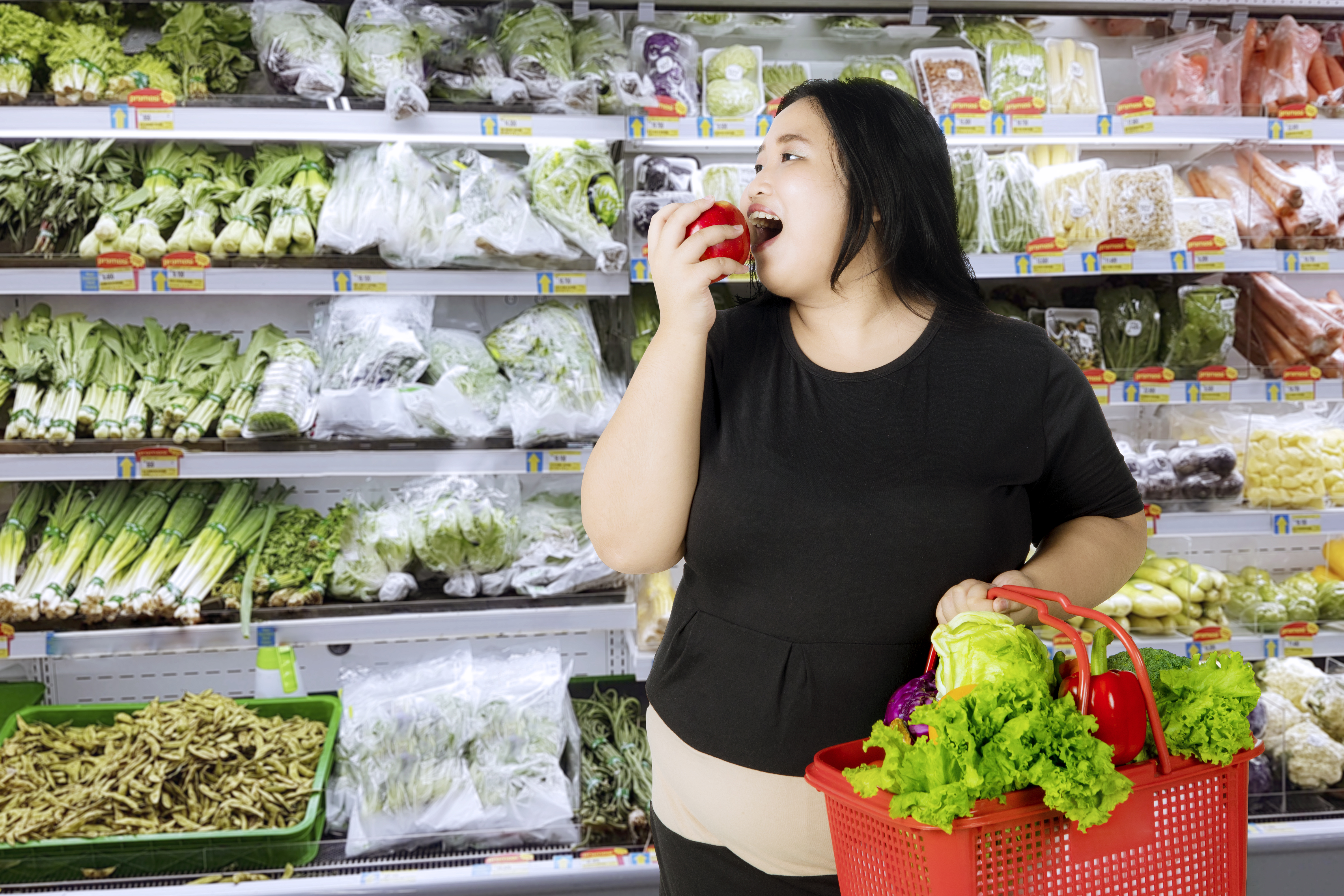 Mujer obesa comiendo una manzana mientras lleva un carrito de compras con verduras orgánicas en el mercado de abarrotes