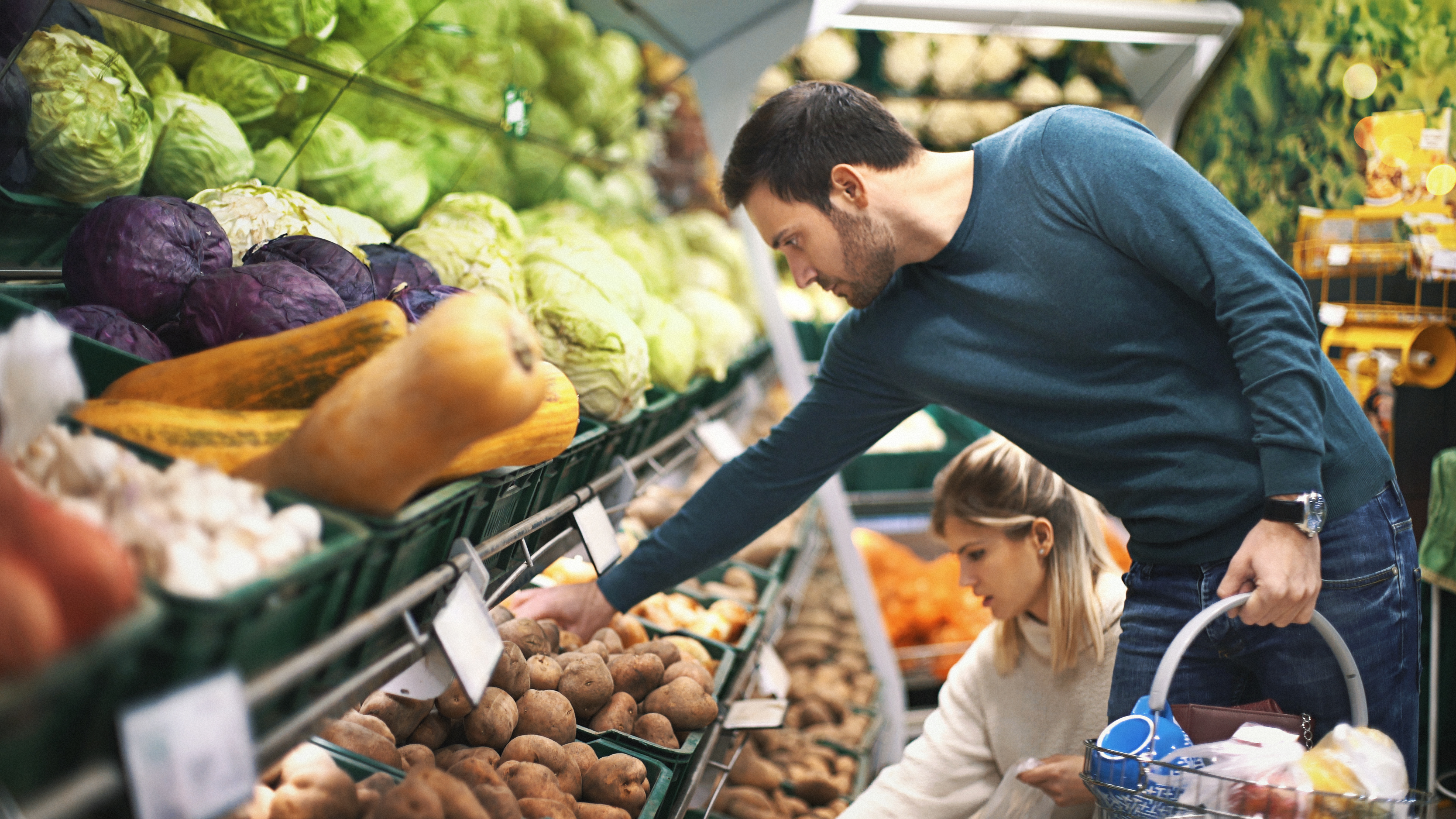 Vista lateral de cerca de una pareja de finales de los años 20 comprando algunas verduras en un supermercado local. Están recogiendo papas y cebollas que necesitarán para la cena. El tipo lleva una cesta de la compra.