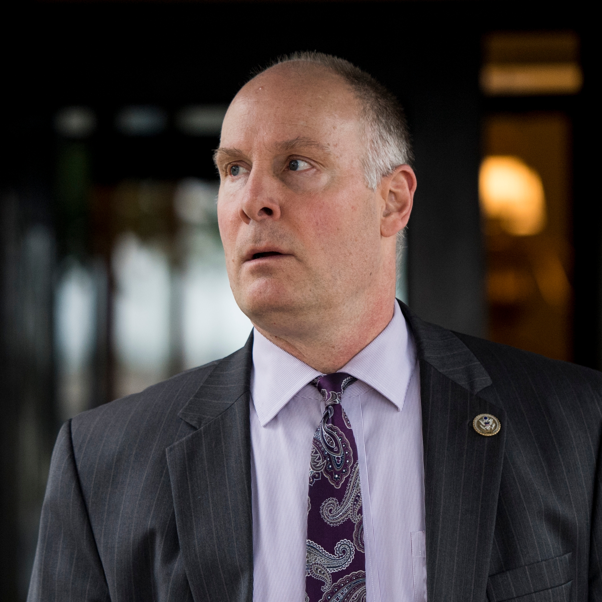 The image shows a middle-aged man with short, thinning hair wearing a dark pinstripe suit jacket over a light purple dress shirt and a purple paisley-patterned tie. He has a slightly open mouth and is looking off to his left with a serious or concerned expression. On his suit jacket's lapel, there is a circular pin, suggesting an official or formal role. The background is blurred with dark tones and some bright, indistinct shapes.