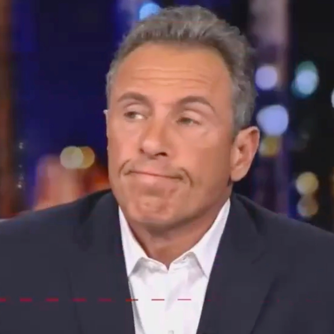 A middle-aged man with short, graying hair is shown in a close-up shot. He is wearing a white dress shirt and a dark jacket, and his facial expression appears to be one of resignation or mild frustration, with his lips pressed together and eyes looking to the side. The background is blurred with various circular lights, suggesting an indoor setting, possibly a studio or nighttime cityscape seen through a window.
