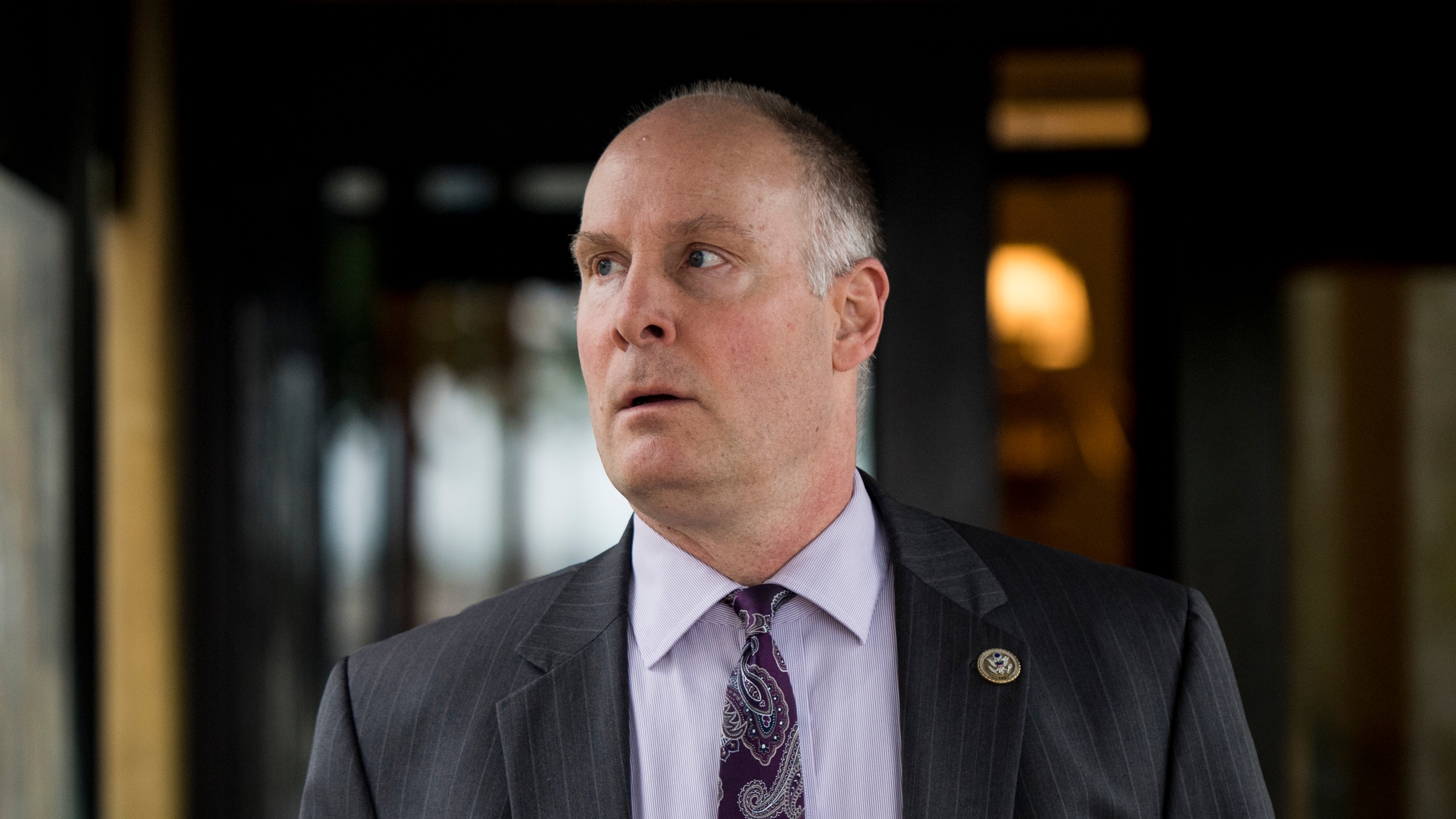 The image shows a middle-aged man with a receding hairline and fair skin, dressed in a dark pinstriped suit jacket, a light lavender dress shirt, and a purple paisley tie. He is looking slightly to his left with a serious or concerned expression on his face. A small pin with a seal is attached to the left lapel of his jacket, indicating some official or governmental affiliation. The background is out of focus, with dark tones and a few blurred lights, suggesting an indoor or semi-indoor setting.
