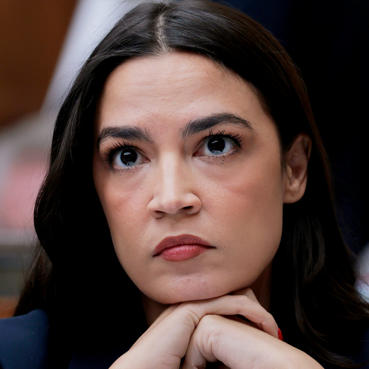 A close-up image shows a woman with long dark hair resting her chin on her clasped hands. Her facial expression is serious and contemplative, with wide eyes looking slightly upward and lips pressed together. The background is out of focus, emphasizing her face and expression.