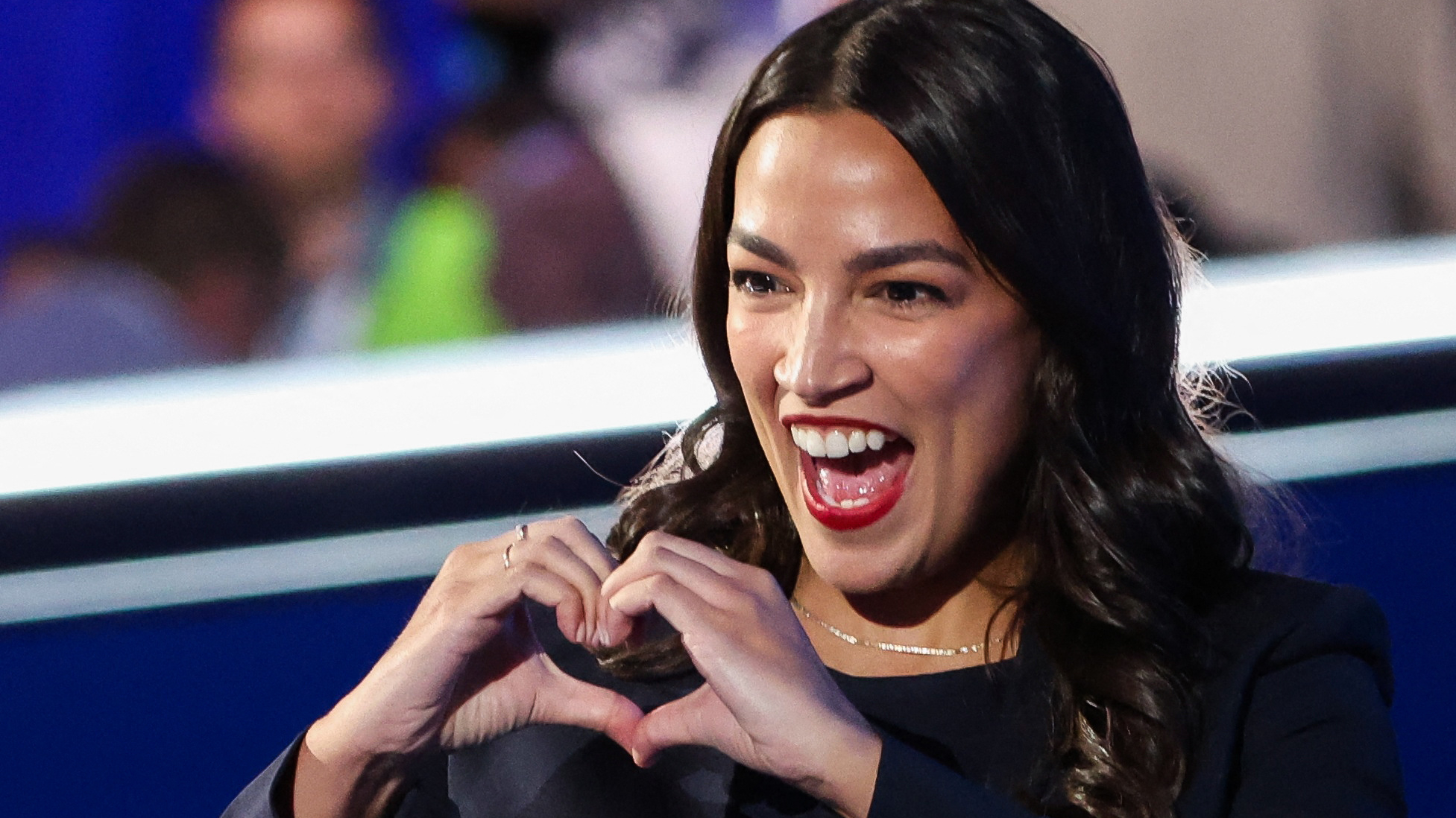 A woman with long dark hair and red lipstick is smiling widely with an open mouth, expressing excitement or happiness. She is forming a heart shape with her hands in front of her chest. The background is blurred, showing indistinct figures and a mix of blue and light colors.