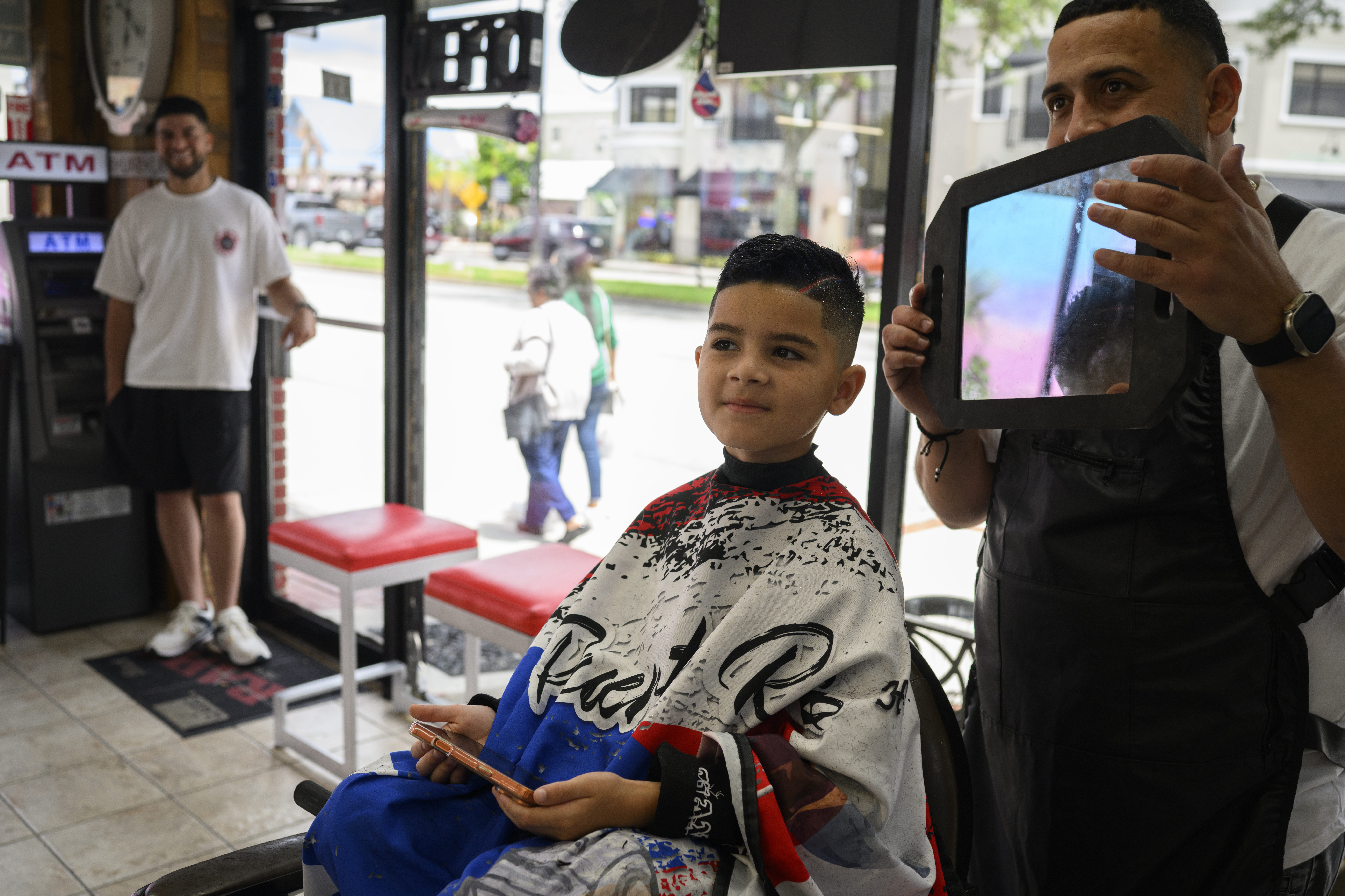 Omar Jose Saldaña, who goes by Ohmie, shows off his work after cutting and coloring the hair of 5-year-old Jahliel Madina, who wears a Puerto Rico-themed cape at MCO Exclusive Barbers in Kissimmee, Florida. (Photos by Thomas Simonetti/For The Washington Post)