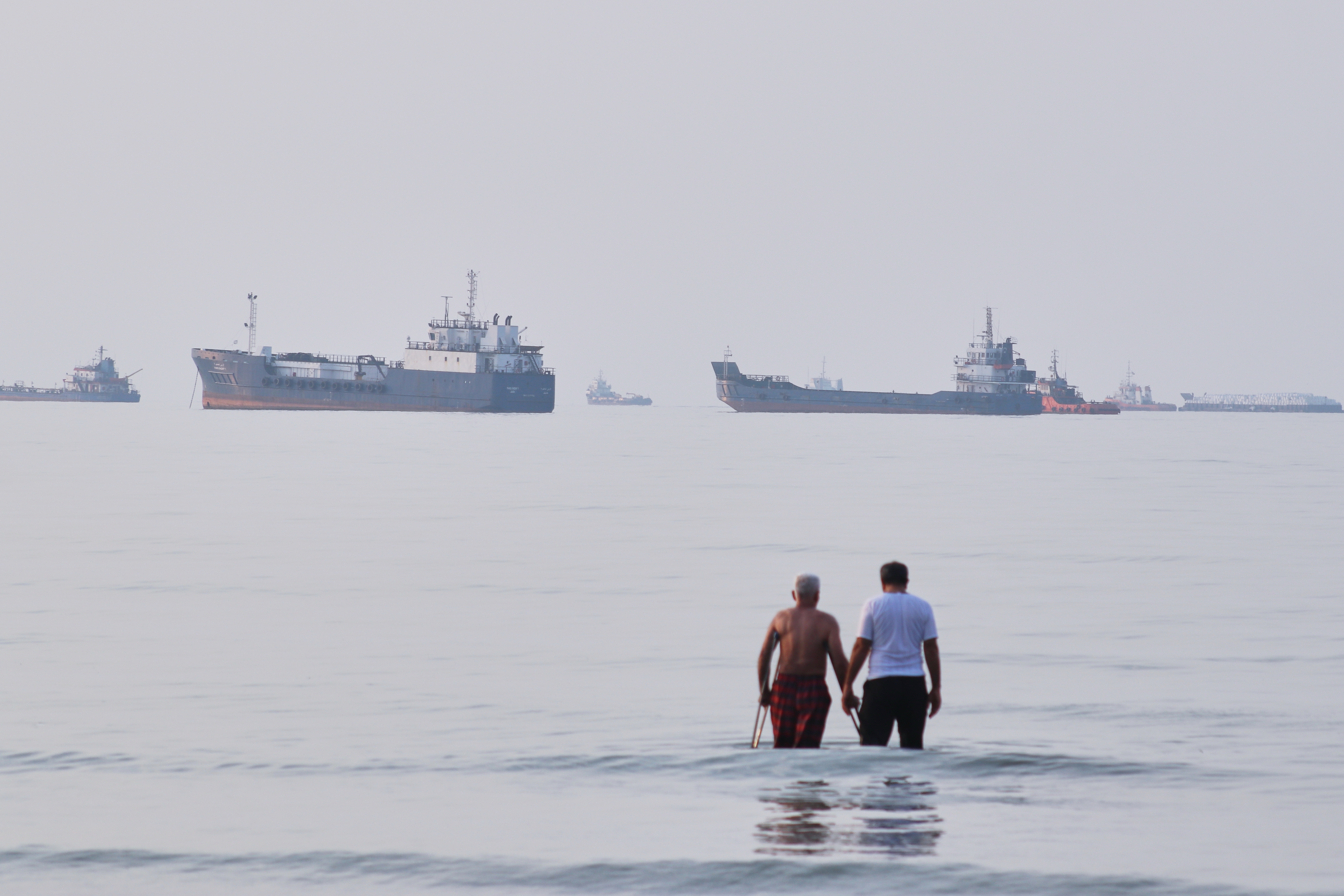 Two men wade in the waters of the Strait of Hormuz on Sunday, with vessels anchored in the background, off Bandar Abbas, Iran. (Razieh Poudat/AP)